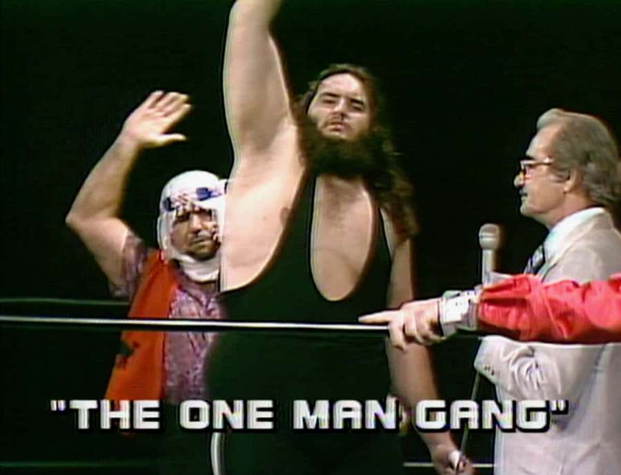 Wrestler raises arm in victory as masked man waves and commentator gestures. — One Man Gang Wallpaper