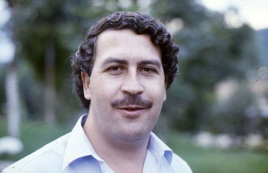 A smiling man with curly hair and mustache in a light shirt outdoors. — Pablo Escobar Picture