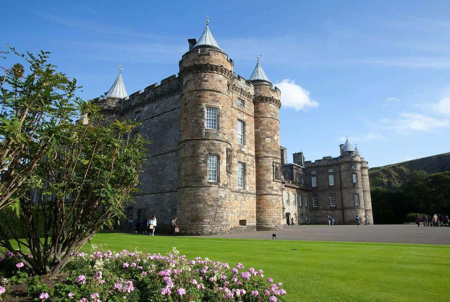 Un majestuoso castillo de piedra con techos azules rodeado de céspedes verdes y flores rosas en un día soleado. — Palacio de Holyrood Fondos de pantalla