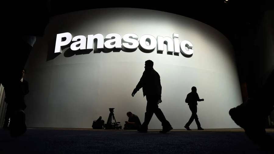 Silhouetten von Menschen, die in einem dunklen Raum an einem großen beleuchteten Panasonic-Logo vorbeigehen. — Panasonic Wallpaper