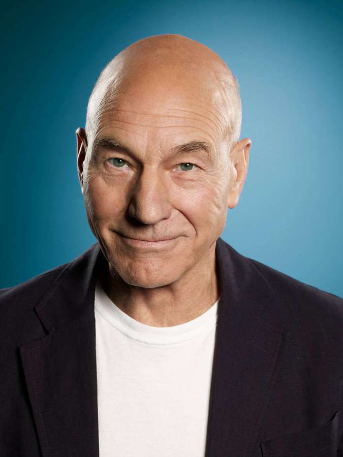 A bald man in a suit smiles gently at the camera against a blue backdrop. — Patrick Stewart Wallpaper