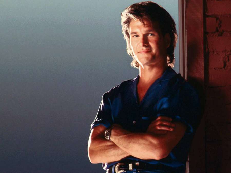 Man in blue shirt leaning against brick wall with arms crossed. — Patrick Swayze Background