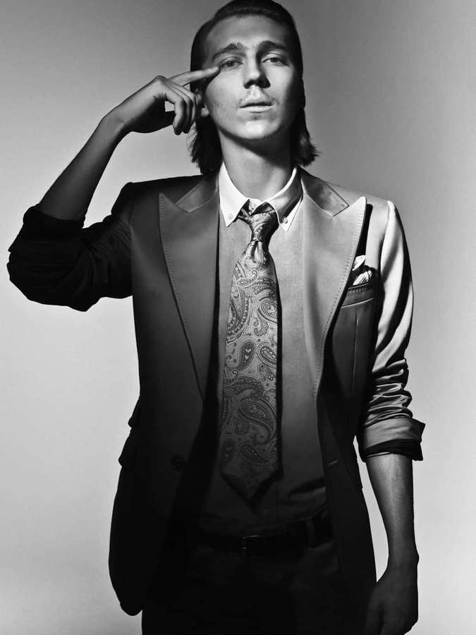 A man in a suit and patterned tie poses with finger to temple in a studio. — Paul Dano Wallpaper