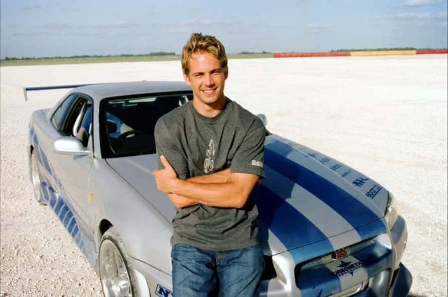 Man in gray t-shirt leaning on silver blue sports car on gravel track — Paul Walker Car Wallpaper