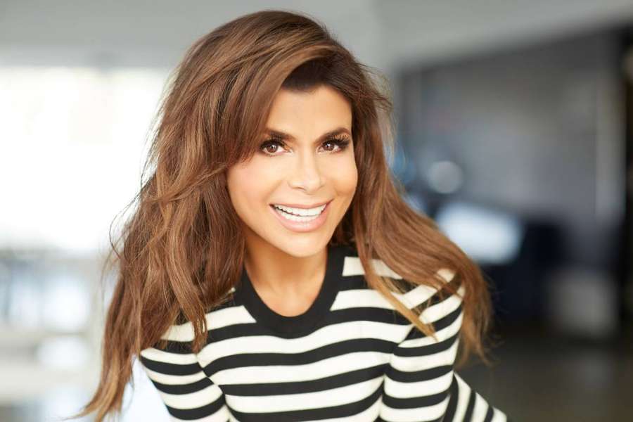 A cheerful woman with long brown hair in a striped top smiles warmly indoors. — Paula Abdul Background