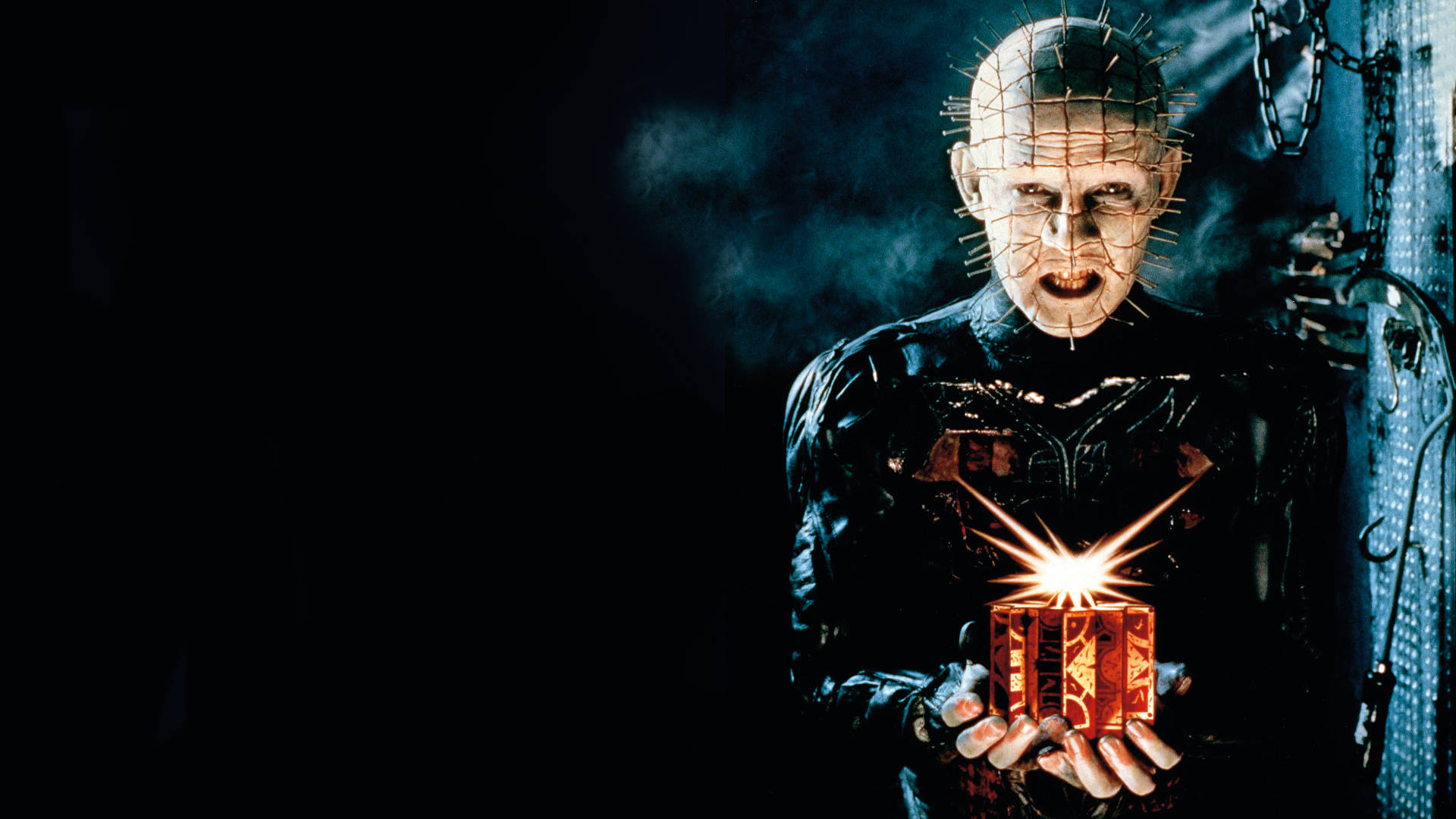 Pinhead holding a glowing puzzle box in a dark industrial setting. — Pinhead Background