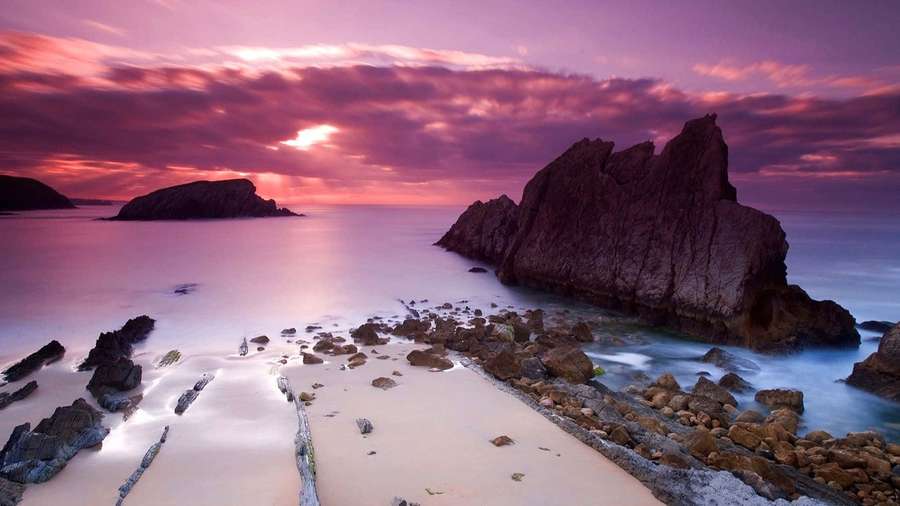 Pink purple sunset over rocky beach with calm water and sand. — Pink Sunset Background