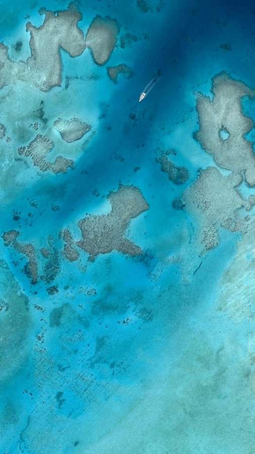 A small boat sails through clear blue waters over a coral reef from above. — Pixel Beach Picture
