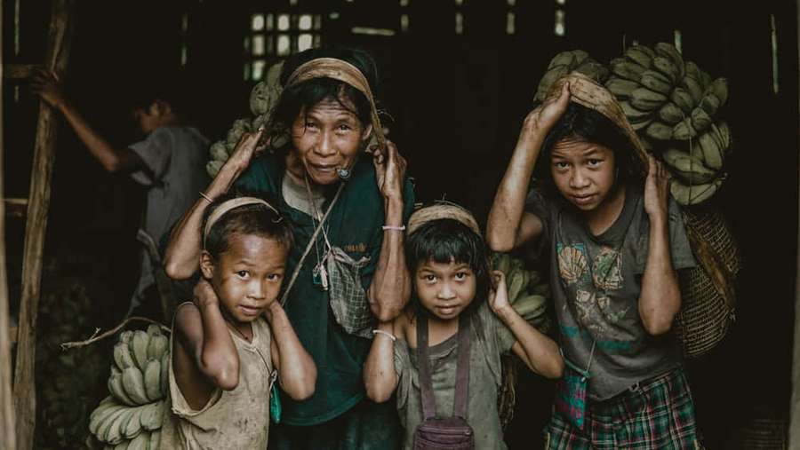 A woman and three children carry banana bunches on their heads indoors. — Poor Wallpaper