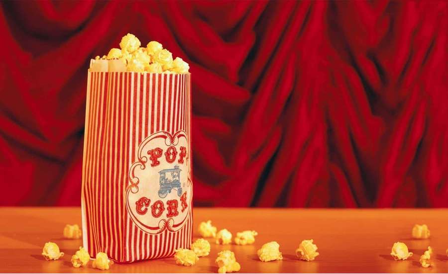 A popcorn bag with spilled kernels on a wooden table in front of a red curtain. — Popcorn Wallpaper
