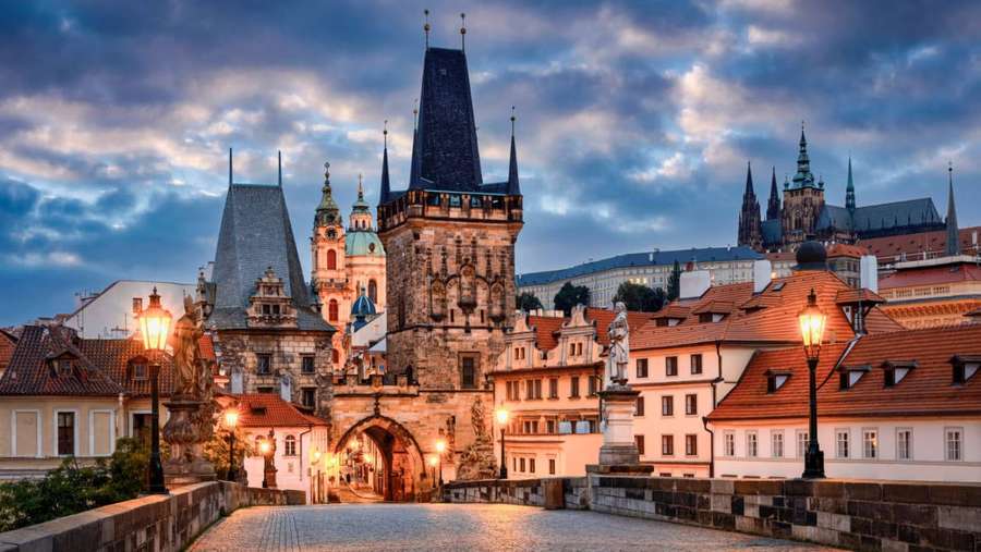 Puente de Carlos en Praga al anochecer con faroles brillantes y agujas góticas bajo un cielo melancólico. — Praga Fondos de pantalla