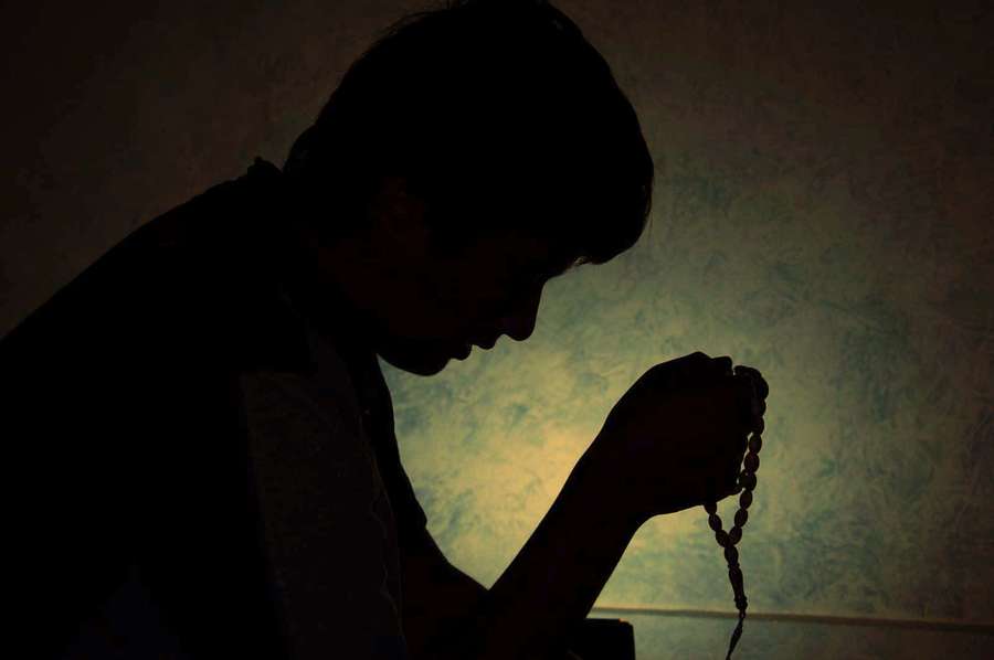 Person in silhouette praying with rosary beads in dim light. — Praying Background