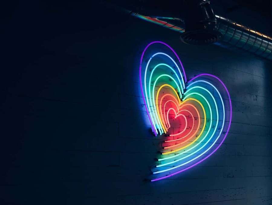 A glowing rainbow heart neon sign on a dark brick wall. — Pride Aesthetic Background