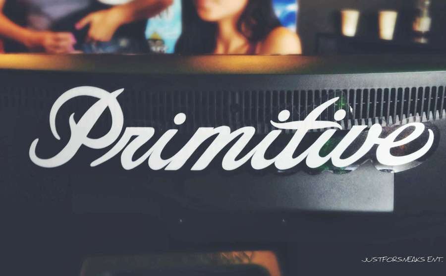 Eine dunkle Bar mit einem weißen kursiven 'Primitive'-Logo und verschwommenen Gästen dahinter. — Primitiv Wallpaper