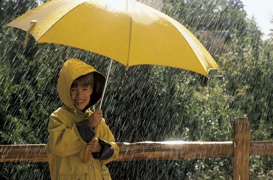 Child in yellow raincoat holding yellow umbrella smiling in rain near wooden fence — Raining Picture