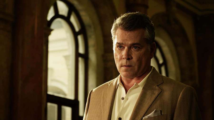 A man in a beige suit stands in a stone hallway with arched windows. — Ray Liotta Wallpaper