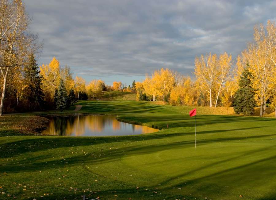 Autumn golf course with green, red flag, pond, and golden trees under cloudy sky. — Red Deer Canada Wallpaper