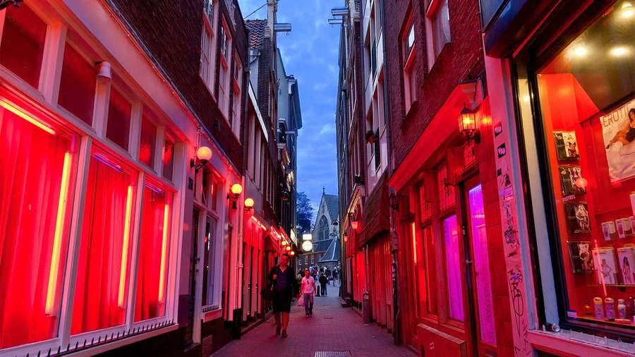 Red neon lights illuminate a narrow city alley at dusk with pedestrians walking. — Red Light District Wallpaper