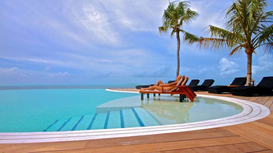 Person lounging by infinity pool with ocean view and palm trees — Resort Wallpaper