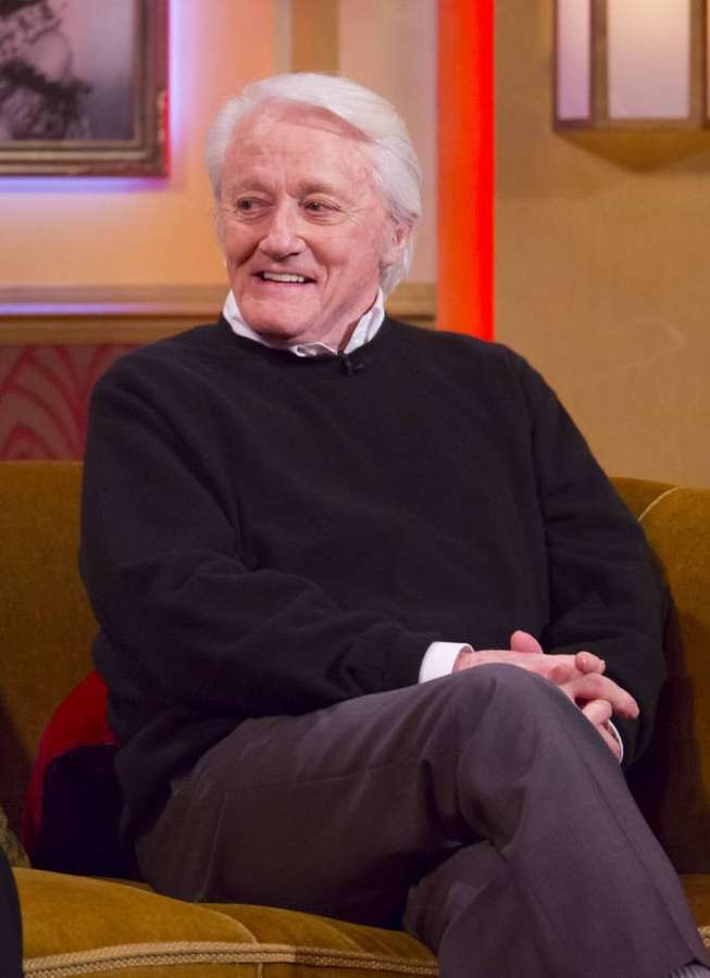 An elderly man with white hair smiles while seated on a mustard yellow couch in a studio. — Robert vaughn Background