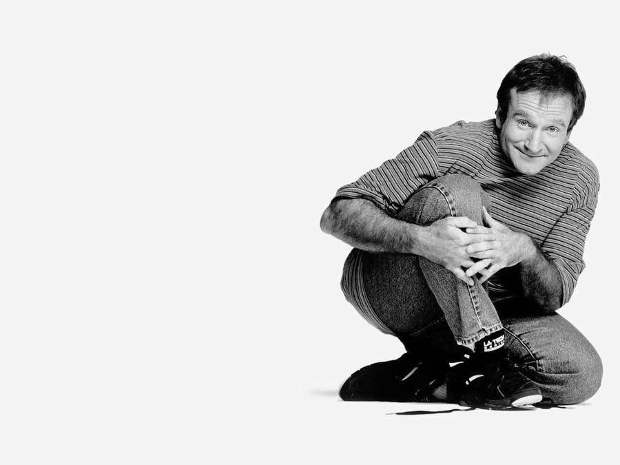 Mann, der in einem Studioporträt mit schlichtem Hintergrund hockt und lächelt. — Robin williams Hintergrundbild