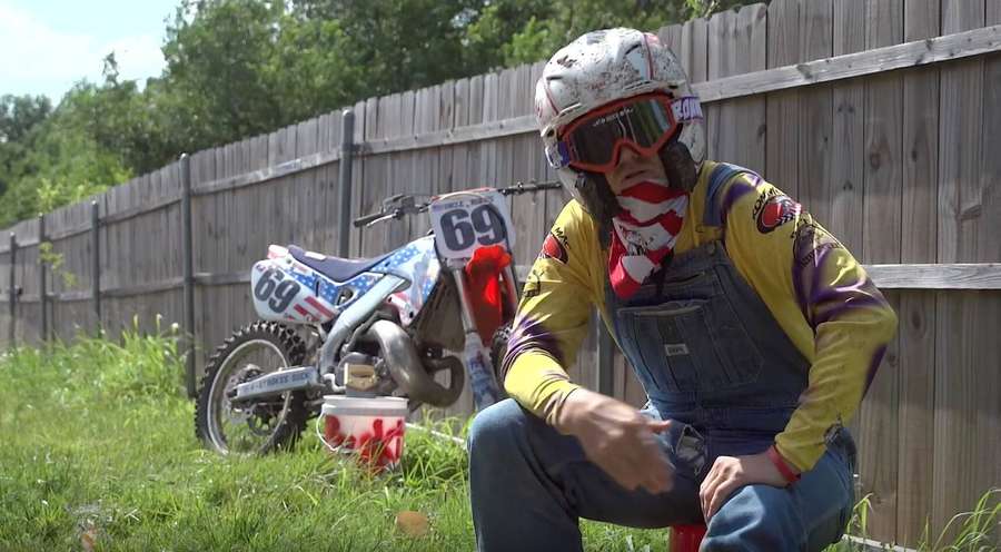 Motocross-Fahrer in patriotischer Kleidung hockt neben seinem Motorrad in der Nähe eines Holzzauns. — Ronnie Mac Bilder