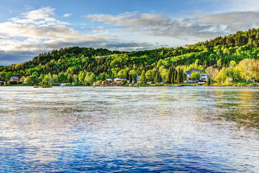 A calm lake with forested hills and houses under a partly cloudy sky. — Saguenay Wallpaper