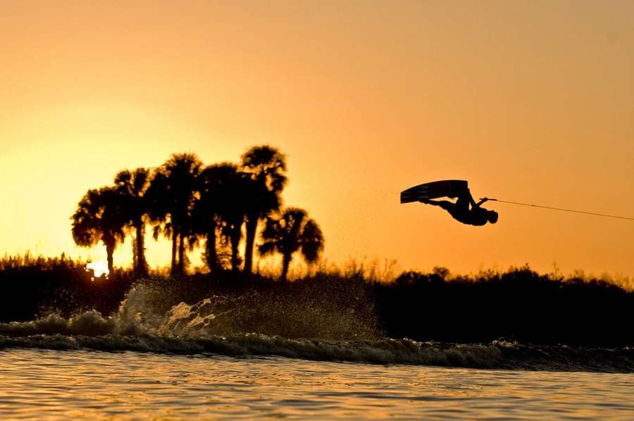 Wakeboarder realizando manobra aérea ao pôr do sol com palmeiras ao fundo. — Saltar Papéis de Parede