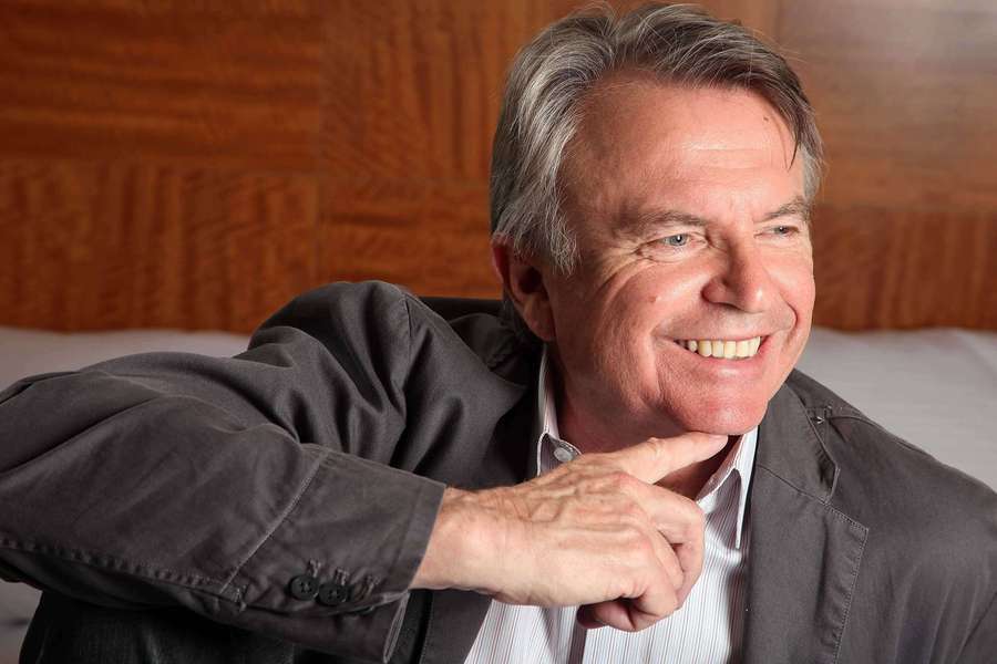 A smiling older man in a jacket resting his chin on his hand indoors. — Sam Neill Wallpaper