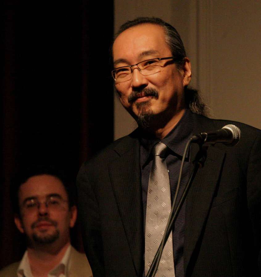 Man in suit speaking into microphone on stage with another man in background. — Satoshi kon Wallpaper