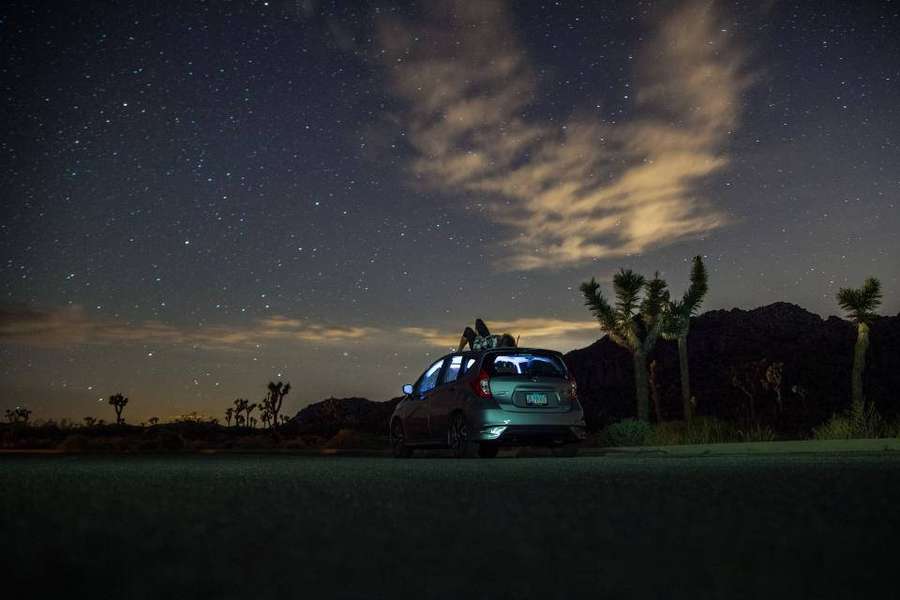 Zwei Personen sitzen auf dem Dach eines silbernen Autos unter einem sternenklaren Wüstenhimmel mit Joshuabäumen. — Schöne Nacht Wallpaper