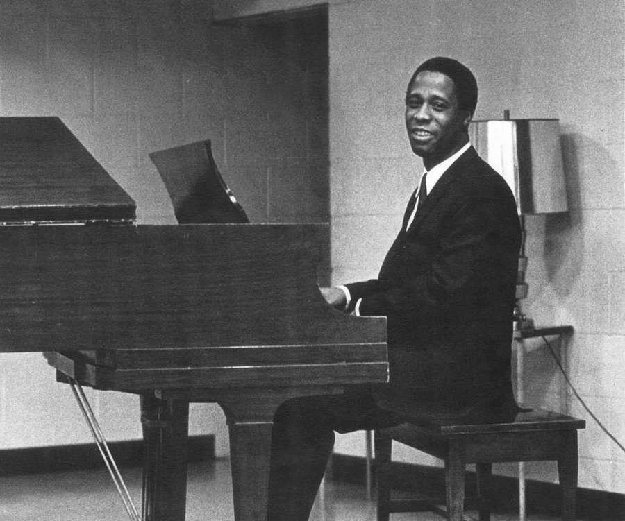 A man in a suit smiles while seated at a grand piano in a room with a brick wall and lamp. — Scott Joplin Wallpaper