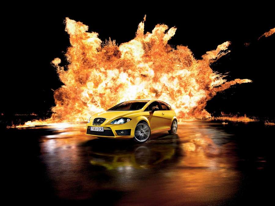 Yellow car speeding past fiery explosion on wet road at night — Seat Wallpaper