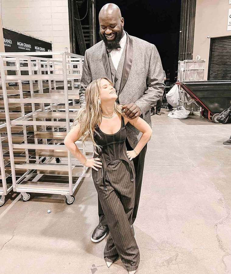 A tall man and woman posing together backstage with metal racks and concrete floor. — Shaq Picture