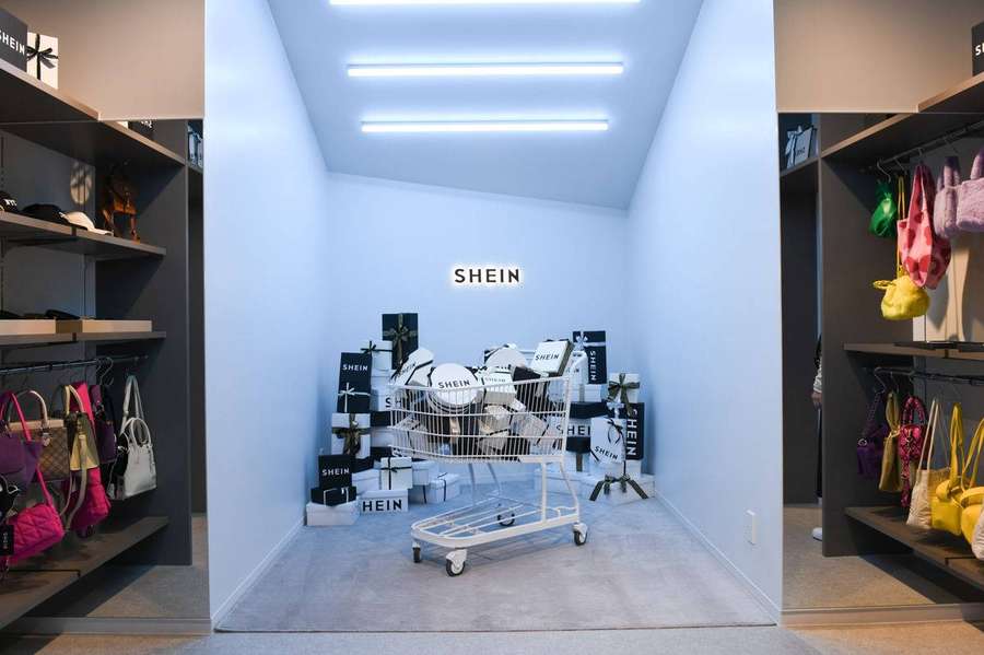 Ein Einkaufswagen voller SHEIN-Kartons in einem modernen Einzelhandelsgeschäft mit Handtaschen in den Regalen. — Shein Bilder