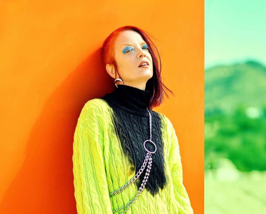 Woman in neon green sweater and silver chain accessories leaning against orange wall with green hills in background. — Shirley Manson Wallpaper