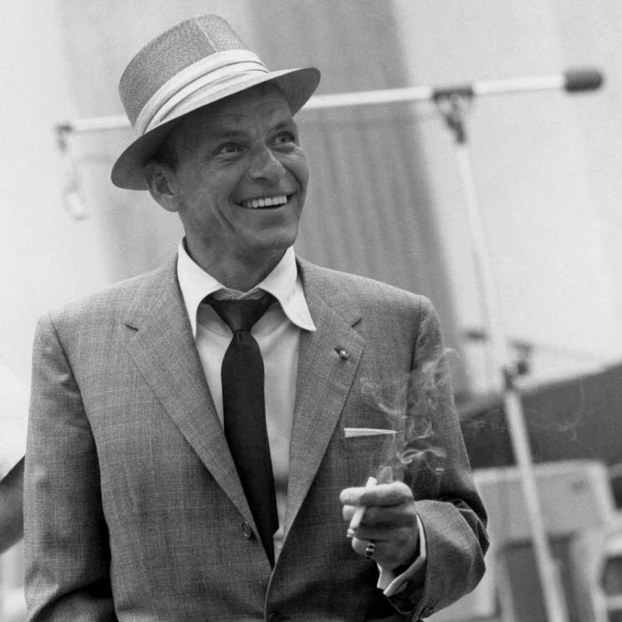 Lächelnder Mann mit Fedora und Anzug, der draußen eine rauchende Zigarette hält. — Sinatra Bilder