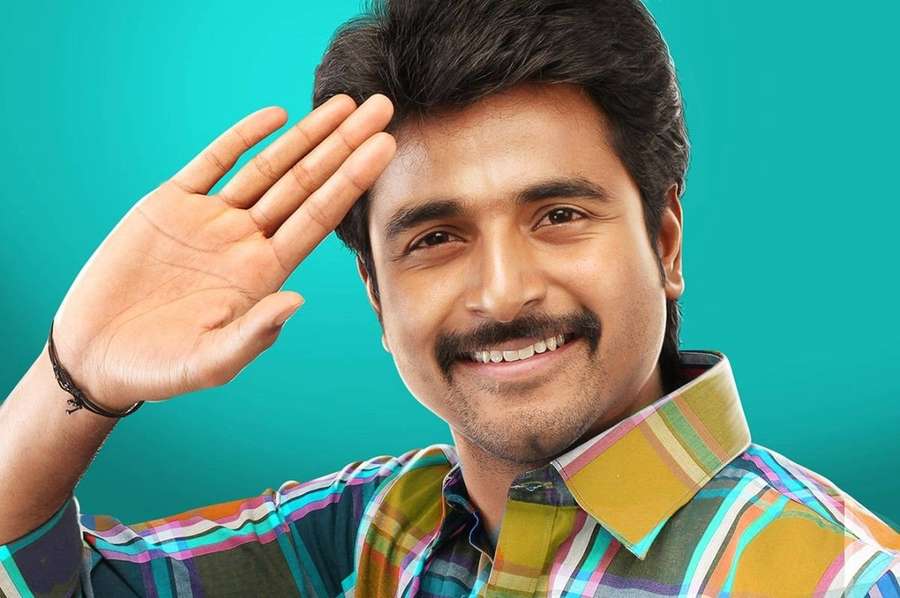 A smiling man in a plaid shirt waves cheerfully against a teal background. — Sivakarthikeyan Wallpaper