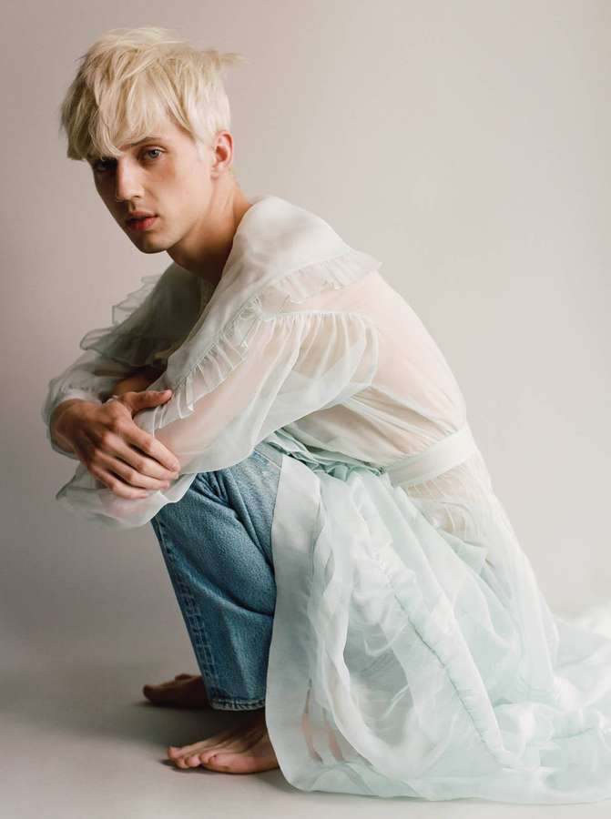 A young blonde man crouches barefoot in jeans and a sheer white ruffled top. — Sivan Background