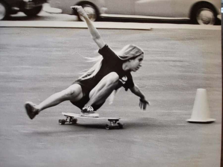 Skatista deslizando baixo passando por um cone no asfalto com carros ao fundo. — Skate retrô Papéis de Parede