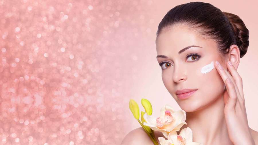 Woman applying skincare cream while holding orchids against glittery pink background. — Skin Background
