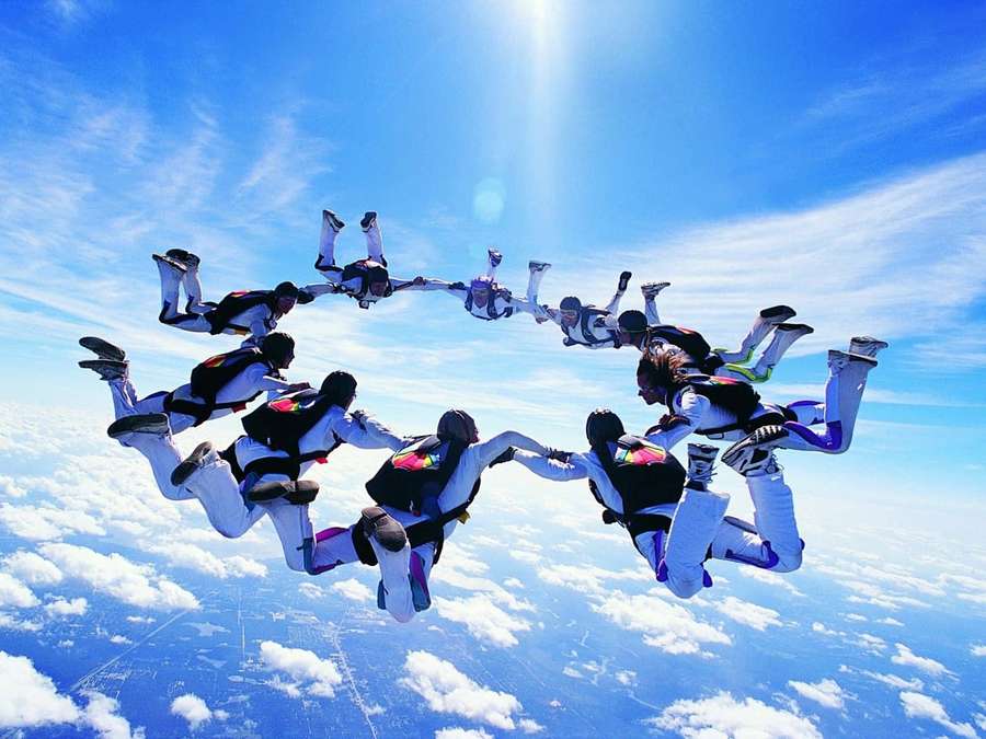 Skydivers holding hands in a circle above clouds under bright sun. — Skydiving Background