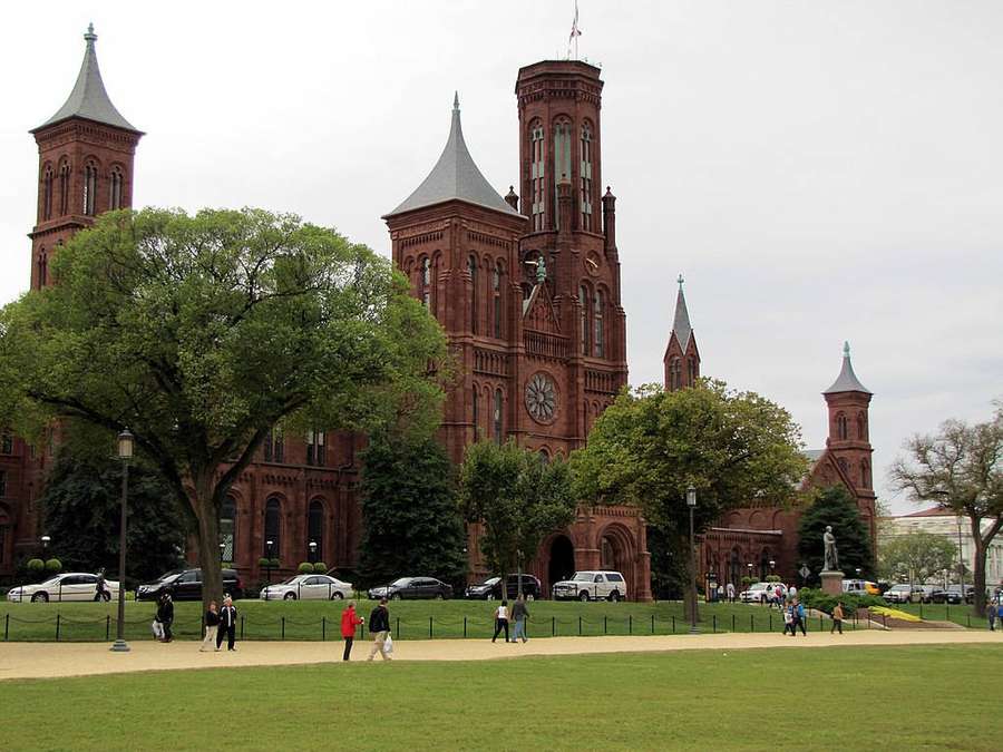 Red brick historic building with towers and clock surrounded by green trees and park visitors. — Smithsonian Wallpaper
