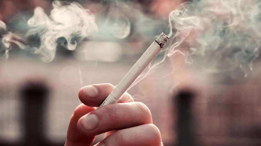 Hand holding a lit cigarette with smoke rising against a blurred city background. — Smoking Background