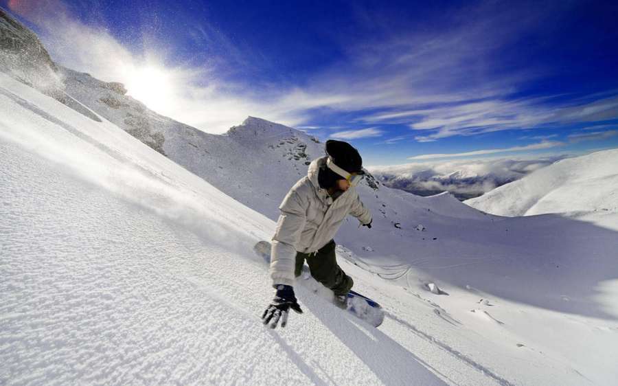 Snowboarder descending a snowy mountain slope with clouds below. — Snowboard Wallpaper