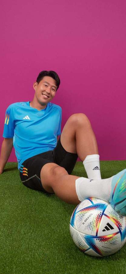Footballer in blue kit smiling while sitting on grass with soccer ball. — Son Wallpaper