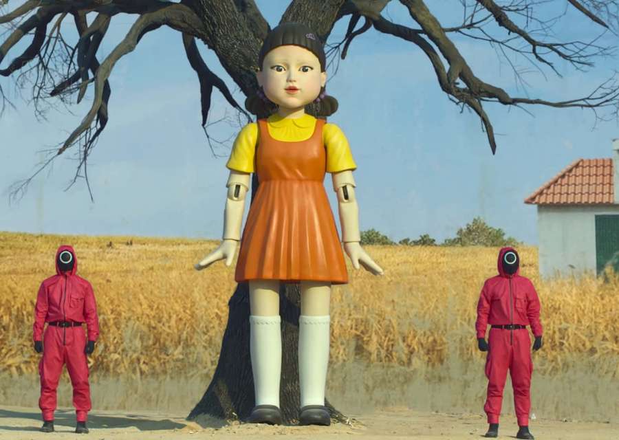 Giant doll under tree with two red-suited figures in field — Squid Game Doll Background
