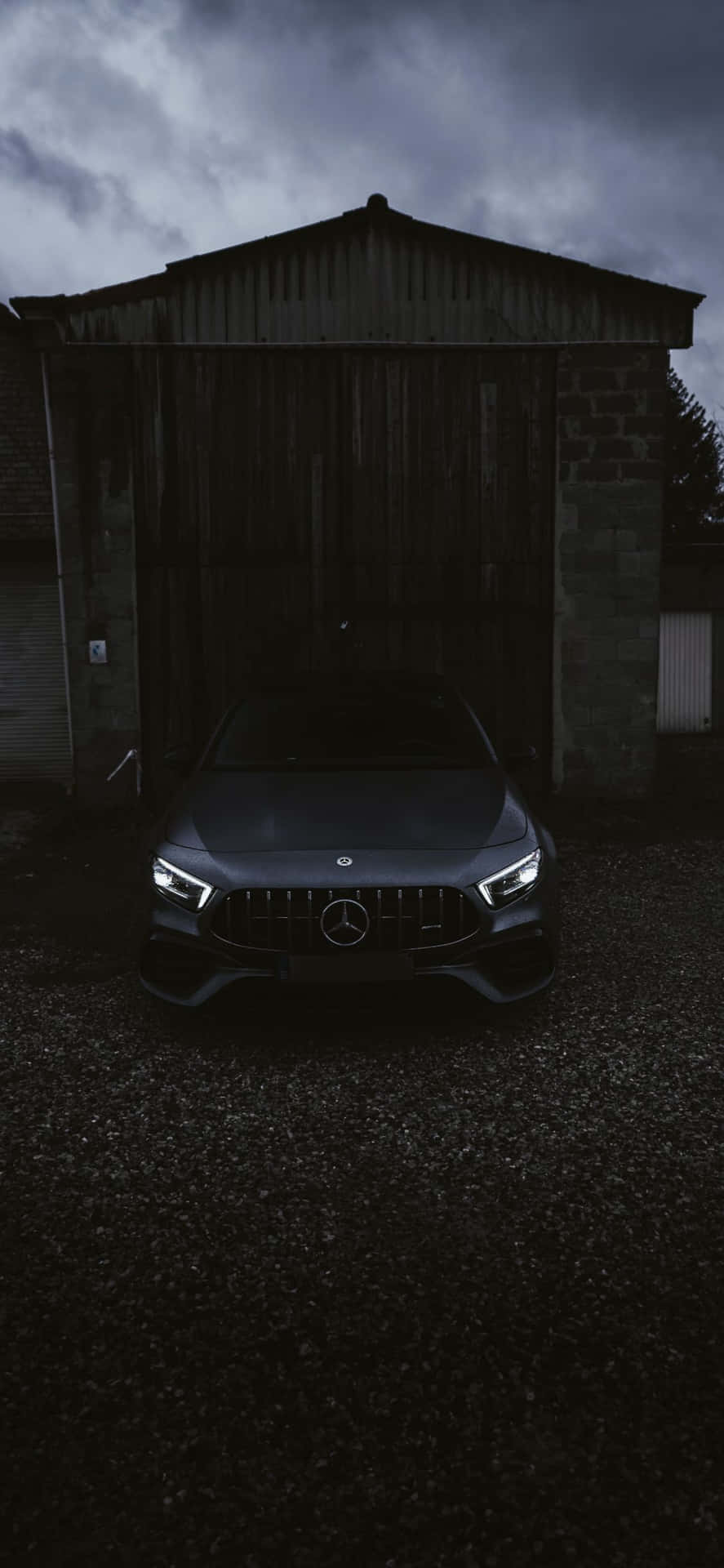 Mercedes-AMG abu-abu gelap diparkir di depan gudang pedesaan di bawah langit yang penuh badai. — Stabil Wallpaper