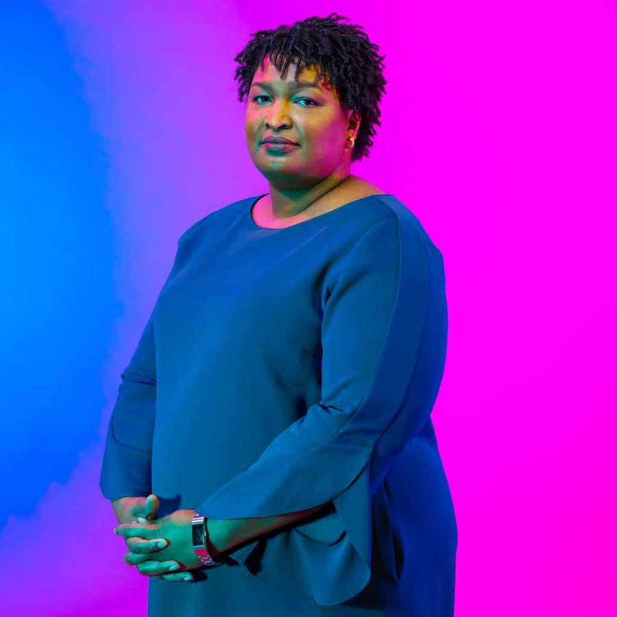 Nainen sinisessä mekossa seisoo itsevarmasti sinistä ja magentaa gradienttitaustaa vasten. — Stacey Abrams Taustakuvat