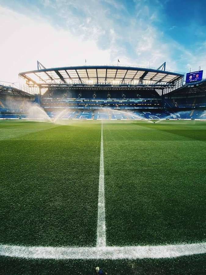 Um estádio de futebol vazio com aspersores regando o campo verde sob um céu ensolarado. — Stamford Papéis de Parede
